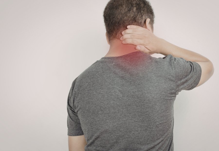 Vista posterior de un hombre tocándose la parte posterior del cuello o nuca, con una zona roja que indica inflamación o dolor muscular.