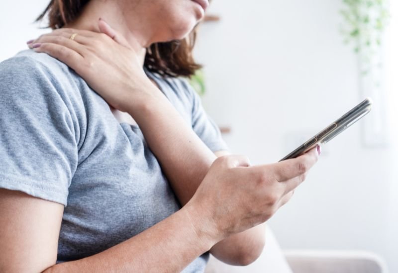 Mujer joven sentada sosteniéndose el hombro con expresión de dolor mientras mira su teléfono móvil de cerca.